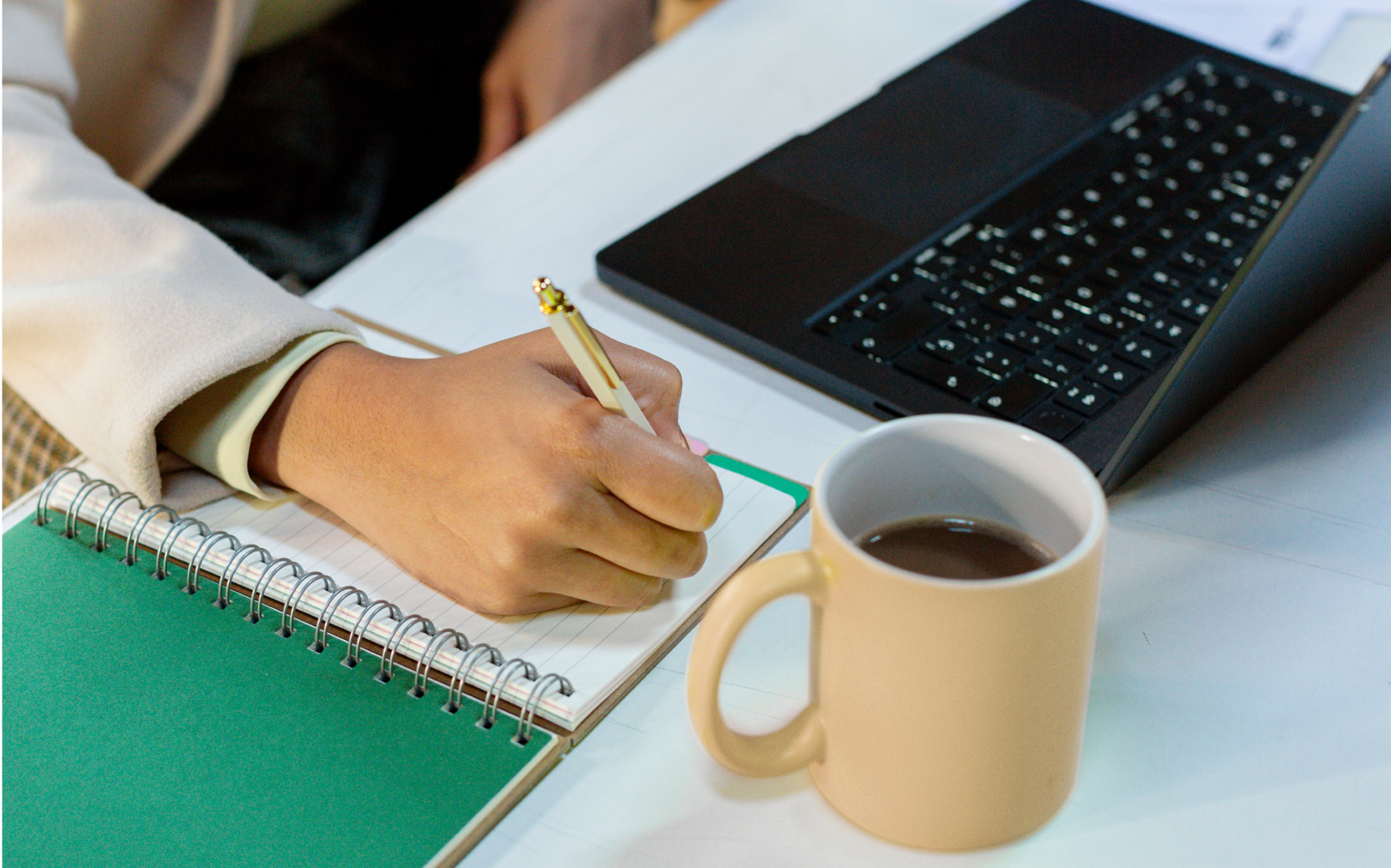 writing in a notebook with coffee and laptop on table